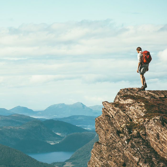 man standing on edge of cliff looking over blue mountains