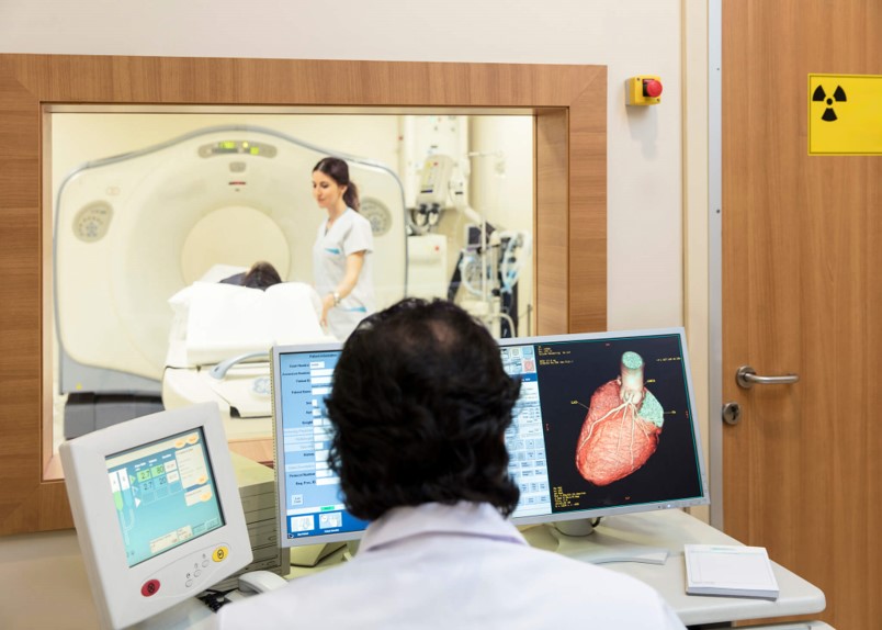 Radiology technologist performing CT scan while cardiologist reviews 3D heart imaging on monitor